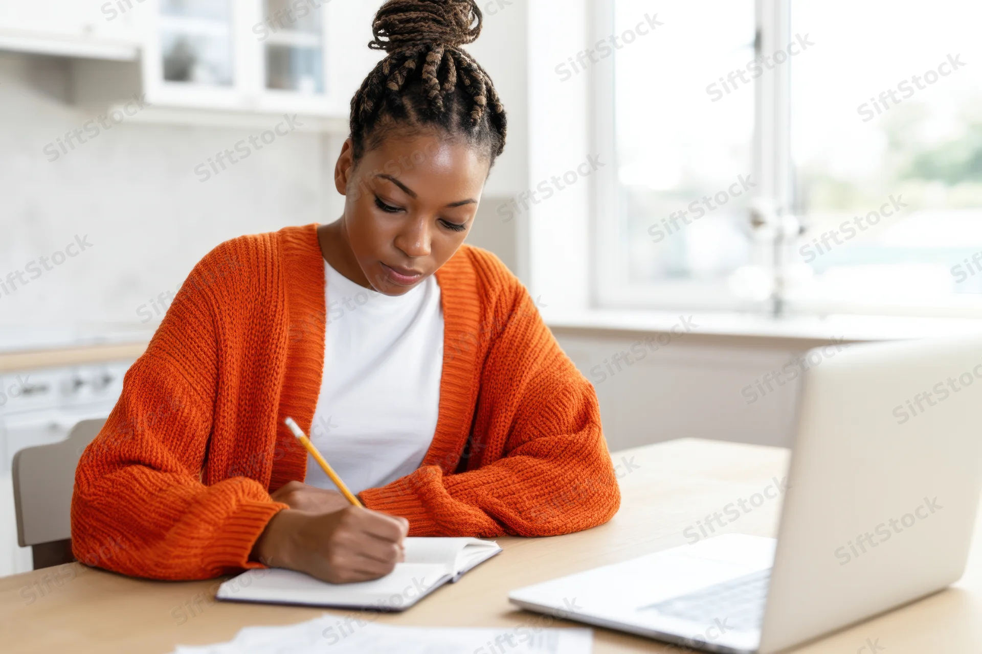 Young woman writing in notebook at kitchen desk with laptop open beside her