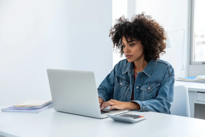 Young woman with curly hair working on laptop at white desk in bright office space