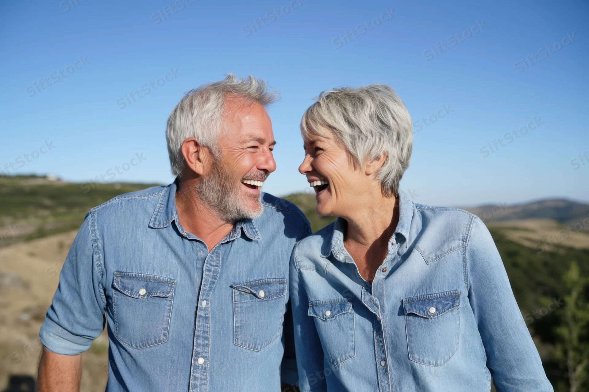 Senior couple laughing together outdoors in denim shirts against a scenic hillside backdrop