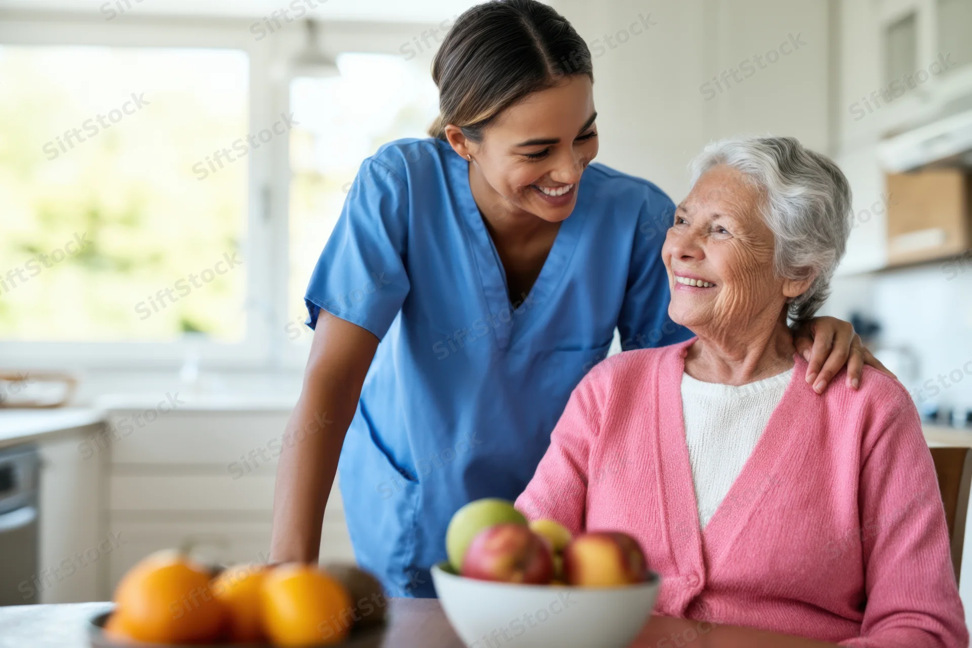 Caregiver smiling at elderly woman seated at kitchen table with fruit bowl