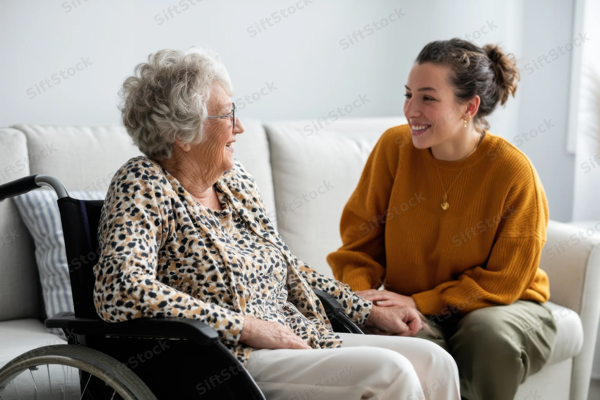 Elderly woman in wheelchair smiling during conversation with caregiver on sofa