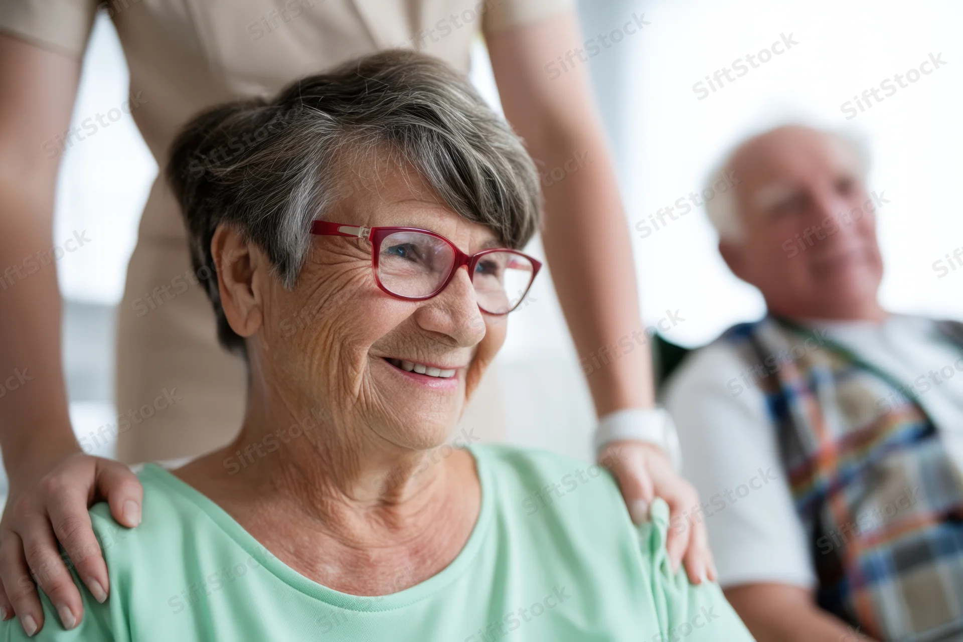 Smiling elderly woman with red-framed glasses receiving shoulder support from a caregiver indoors