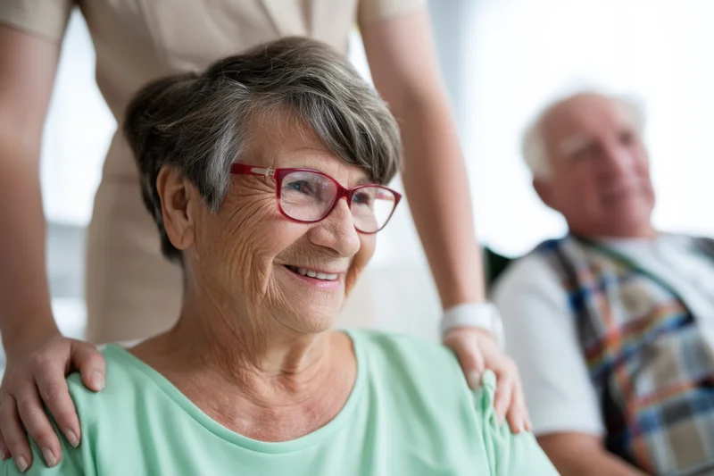 Smiling elderly woman with red-framed glasses receiving shoulder support from a caregiver indoors