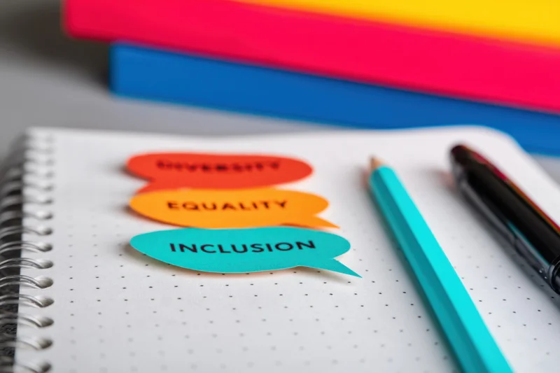 Colorful speech bubble notes with diversity equality inclusion text on a dotted notebook page with pens and pencils nearby