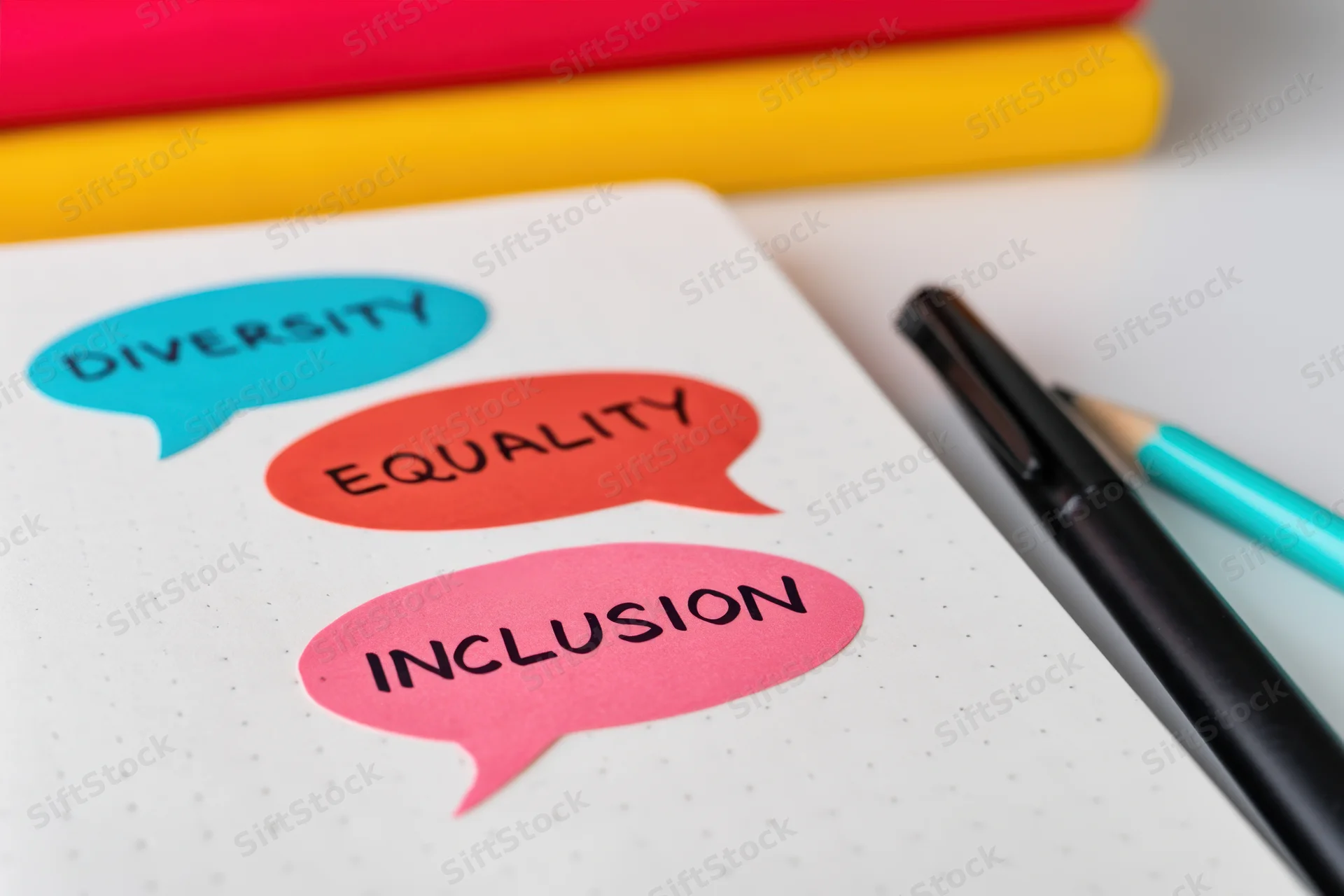 Colorful speech bubbles with diversity equality inclusion text on a notebook beside pens and pencils on a desk