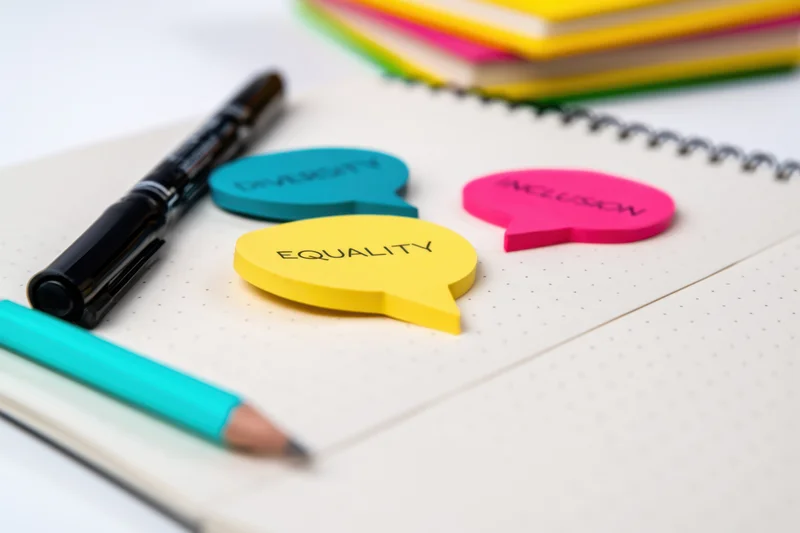 Colorful speech bubble notes with diversity equality inclusion text on a notebook with pens and pencils in an office workspace