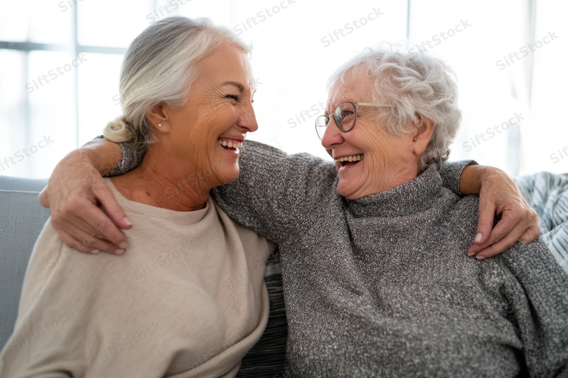 Two elderly women laughing and embracing on a couch in a bright living room