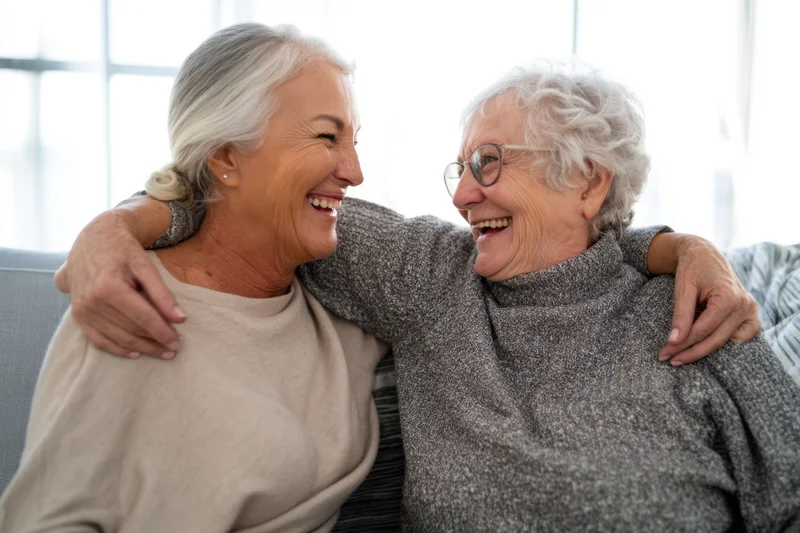 Two elderly women laughing and embracing on a couch in a bright living room