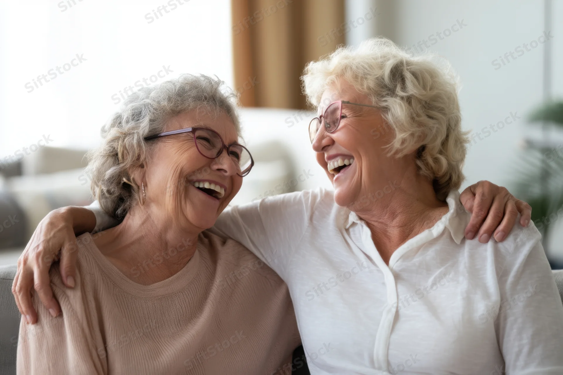 Two elderly women laughing together with arms around each other on a couch indoors