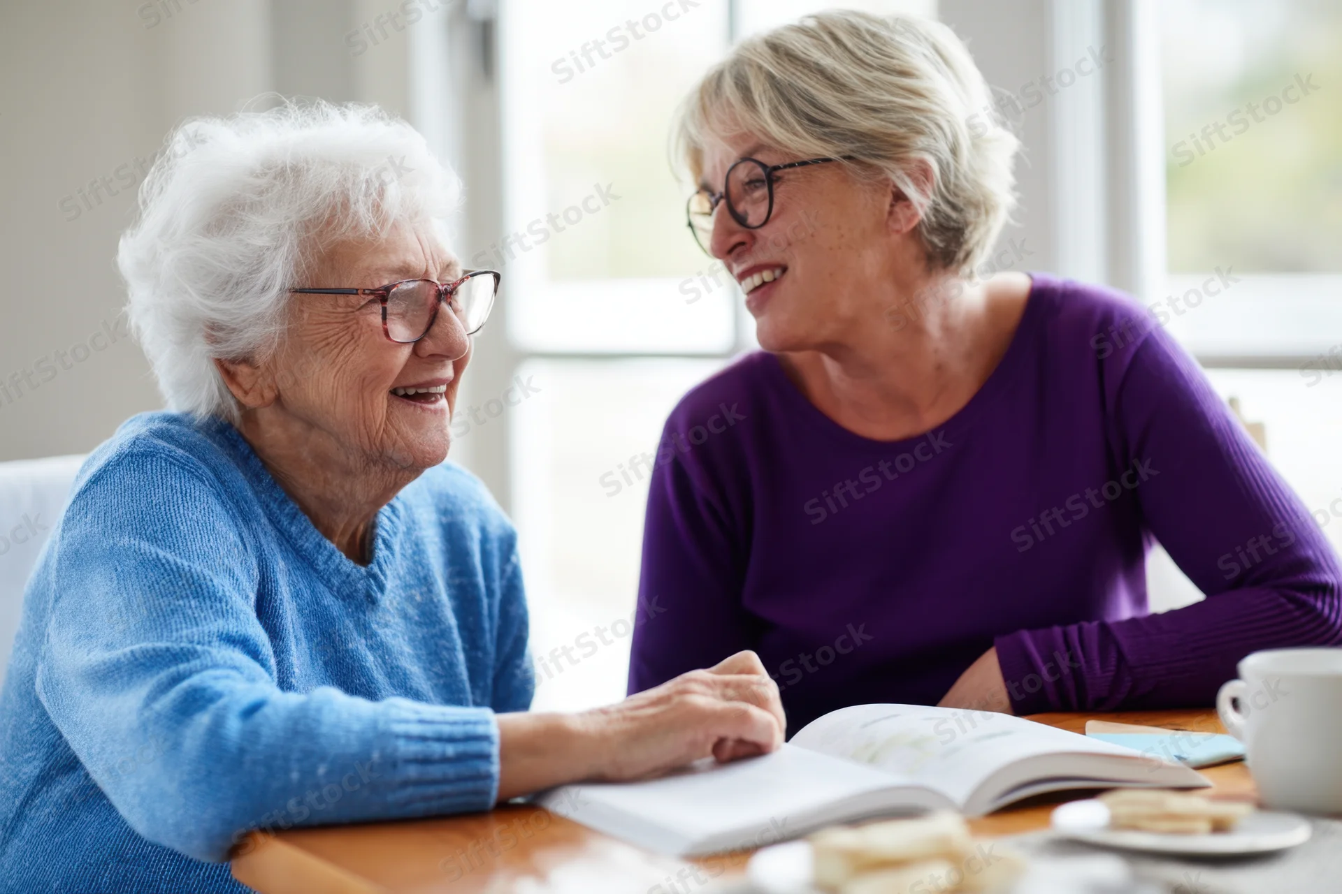 Two women smiling and talking at a kitchen table with a book and coffee