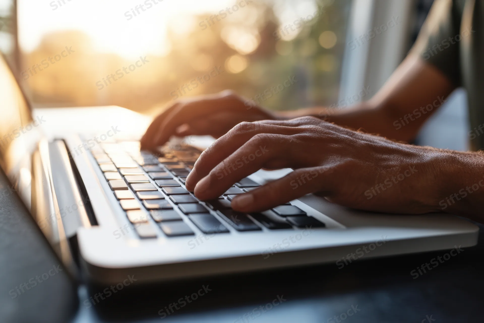 Hands typing on a laptop keyboard on a dark desk near a sunlit window
