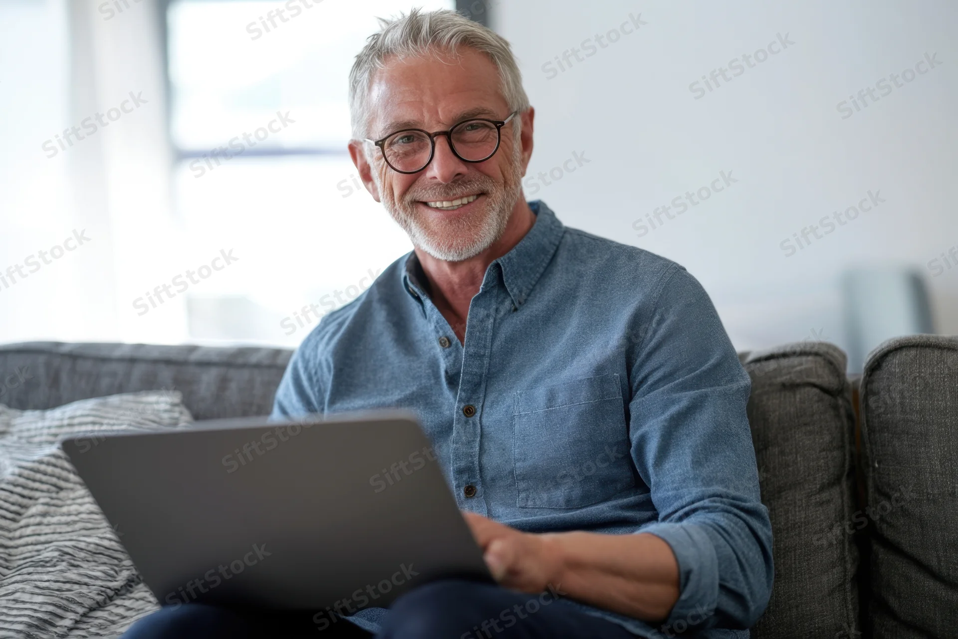 Smiling middle-aged man with gray hair and glasses sitting on a couch using a laptop at home