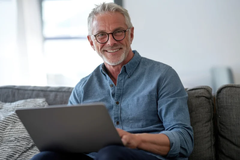 Smiling middle-aged man with gray hair and glasses sitting on a couch using a laptop at home