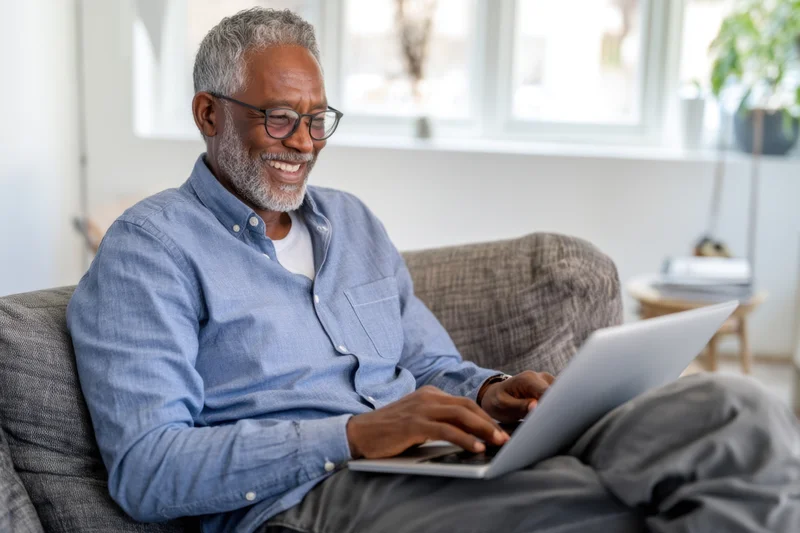 Smiling senior man typing on laptop while sitting on couch at home