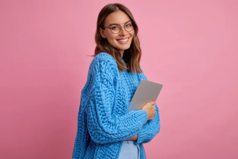 Smiling woman in chunky blue knit sweater holding tablet against pink background