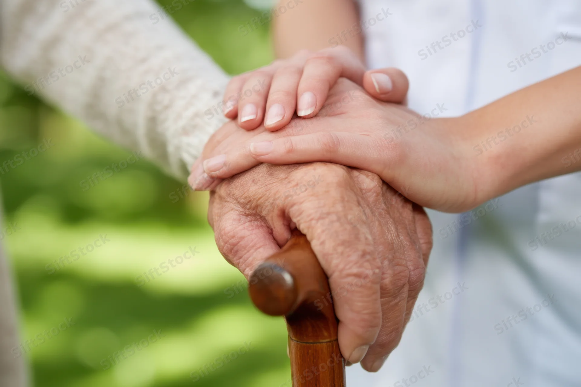 Elderly hand gripping a wooden cane while a younger caregiver’s hand rests gently on top in a garden setting