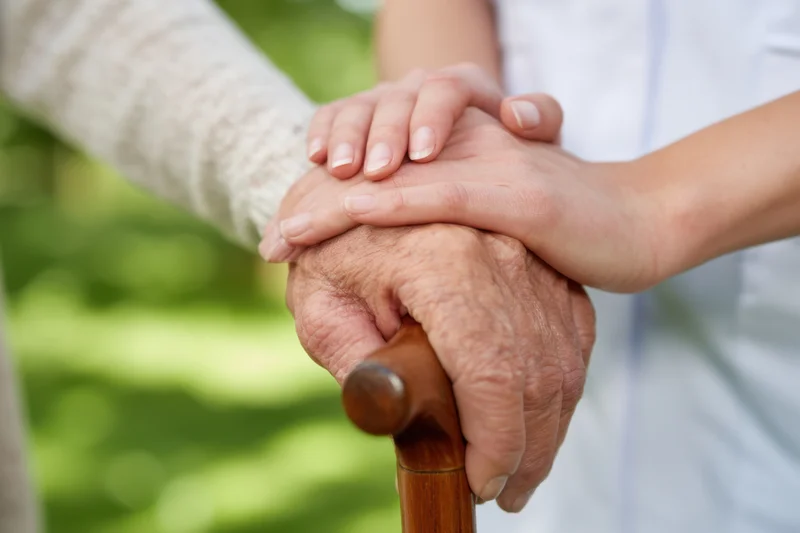 Elderly hand gripping a wooden cane while a younger caregiver’s hand rests gently on top in a garden setting