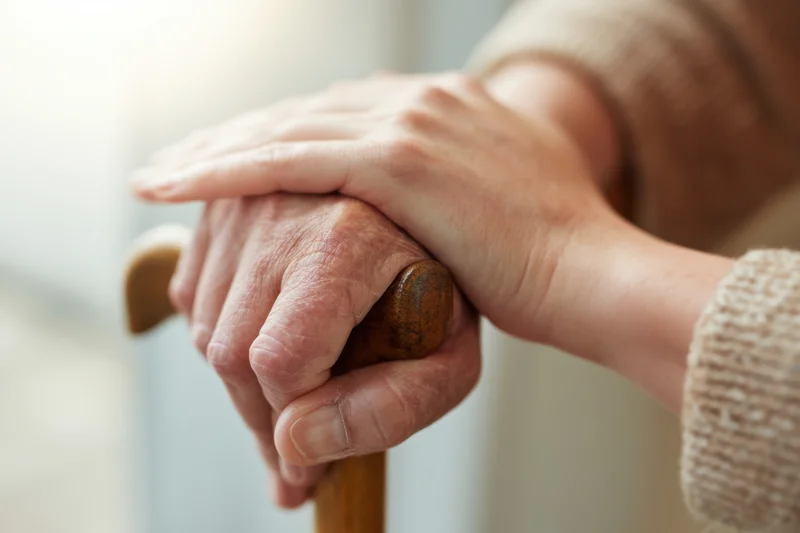 Elderly hand gripping a wooden cane, gently held by a younger hand in a moment of support