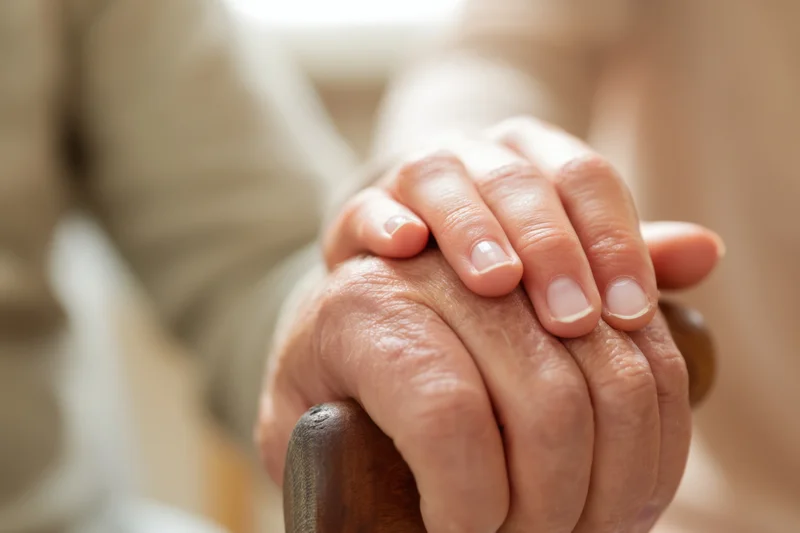 Elderly hand gripping a wooden cane with a younger hand resting on top for support