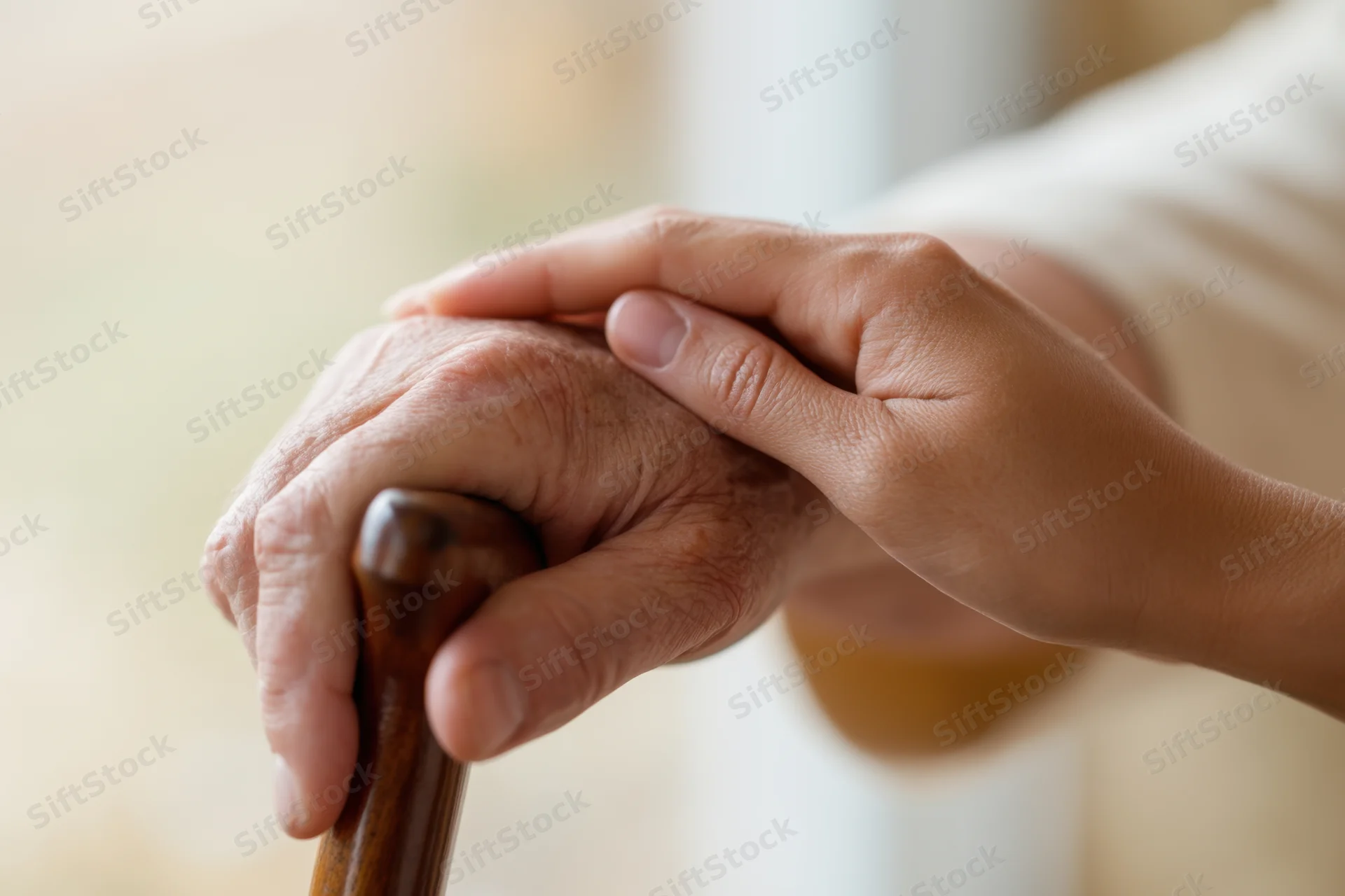 Elderly hand gripping wooden cane, gently held by another hand in a supportive gesture indoors