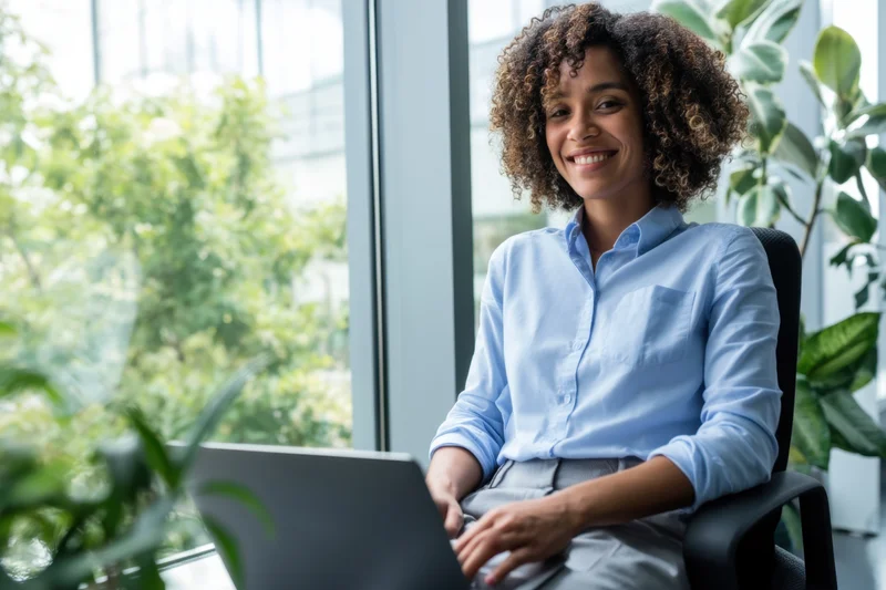 Woman smiling while using laptop at office desk by large window
