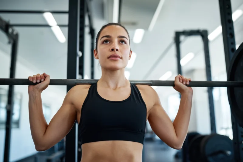Woman holding barbell across shoulders in gym, focused expression, wearing black sports bra