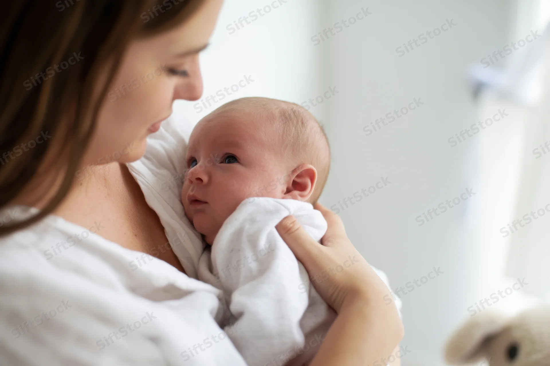 Mother cradling newborn baby wrapped in white blanket in bright room