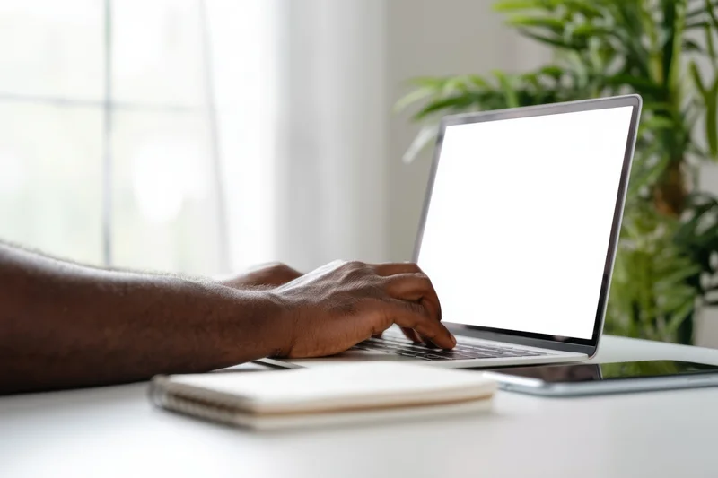 Man typing on a slim laptop with blank white screen at a desk near a window