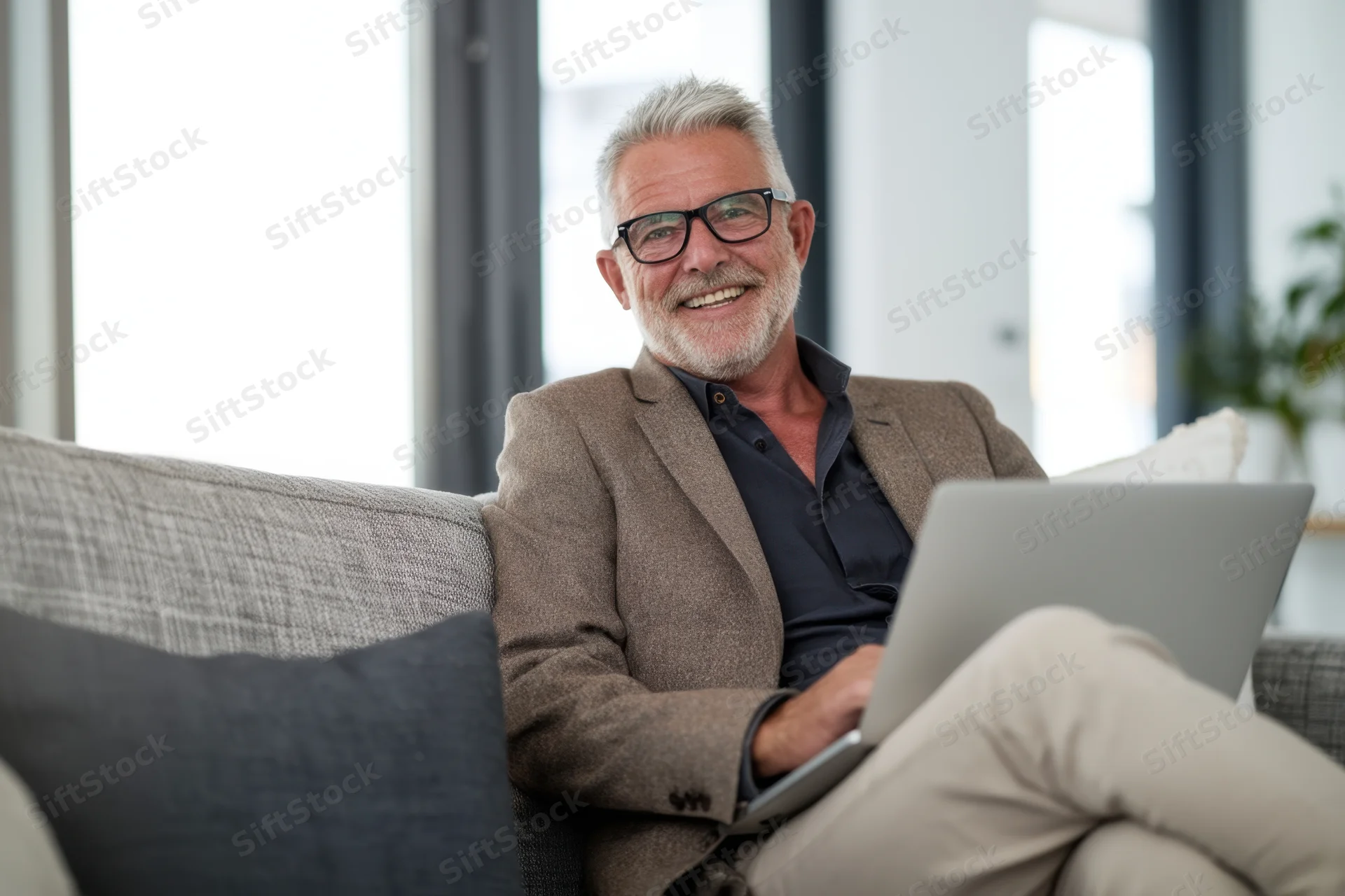 Smiling middle-aged man with gray hair and glasses using laptop on couch at home