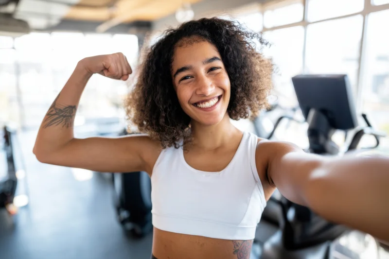 Young woman flexing bicep and smiling while taking selfie in gym
