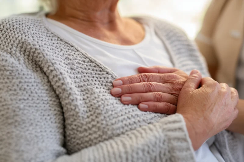 Elderly woman with hands placed gently on her chest, wearing a light gray knitted cardigan over a white shirt, indoors