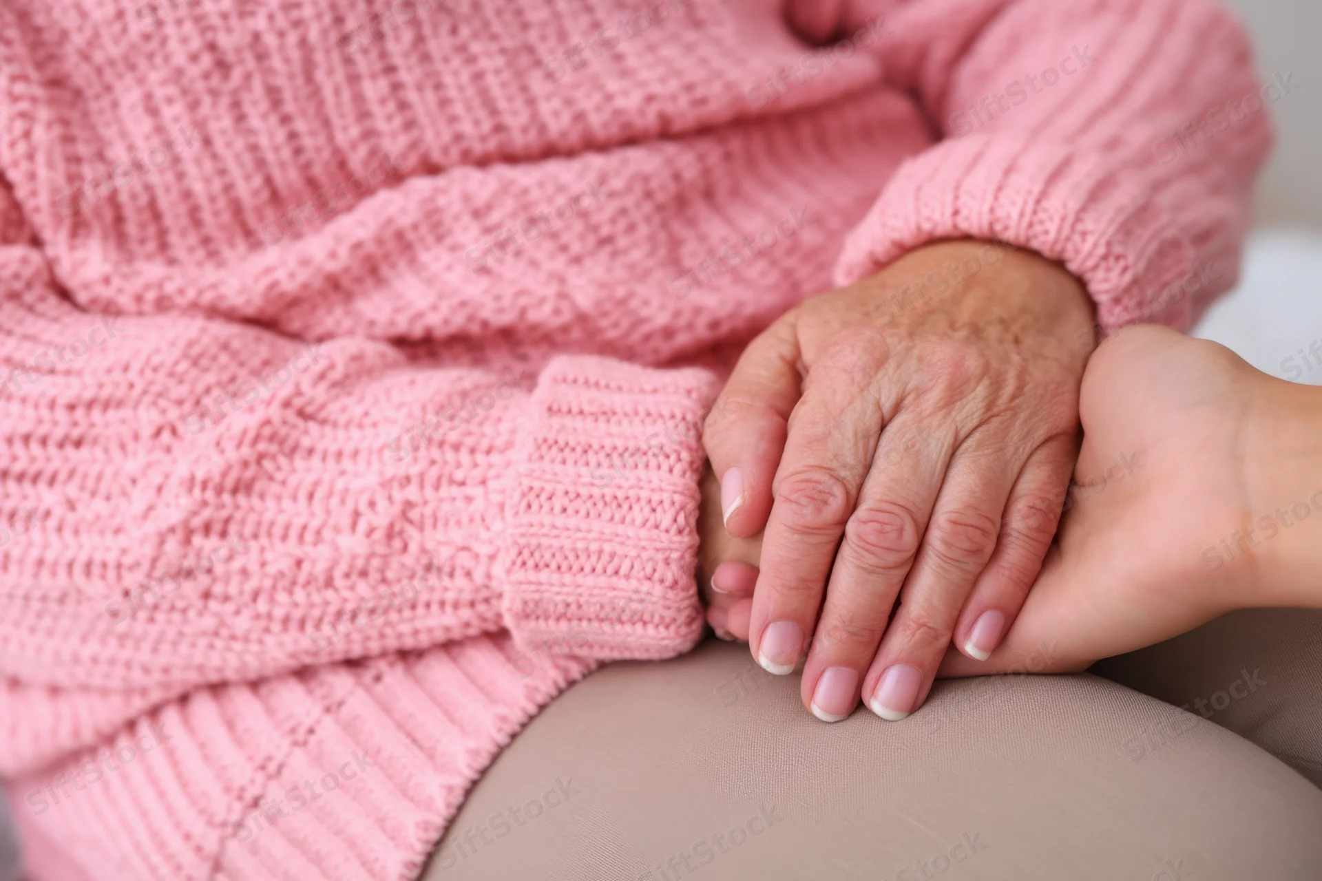 Elderly woman in pink knitted sweater holding hands with a younger person's hand on her lap