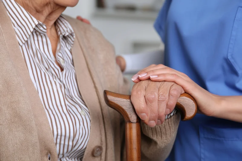Elderly woman holding a wooden cane while a caregiver in blue scrubs supports her hand