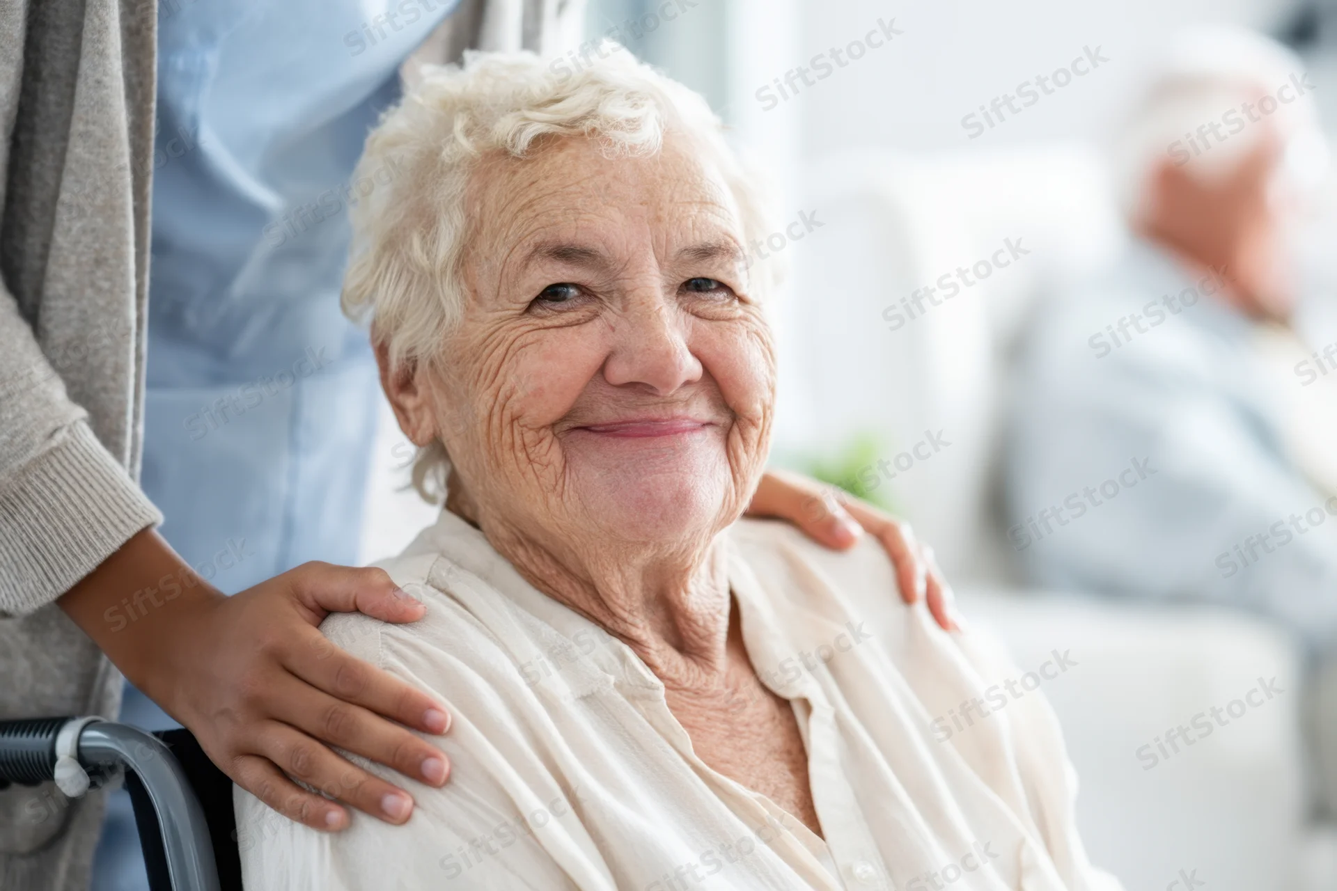 Elderly woman smiling in wheelchair with caregiver’s hands on her shoulders indoors