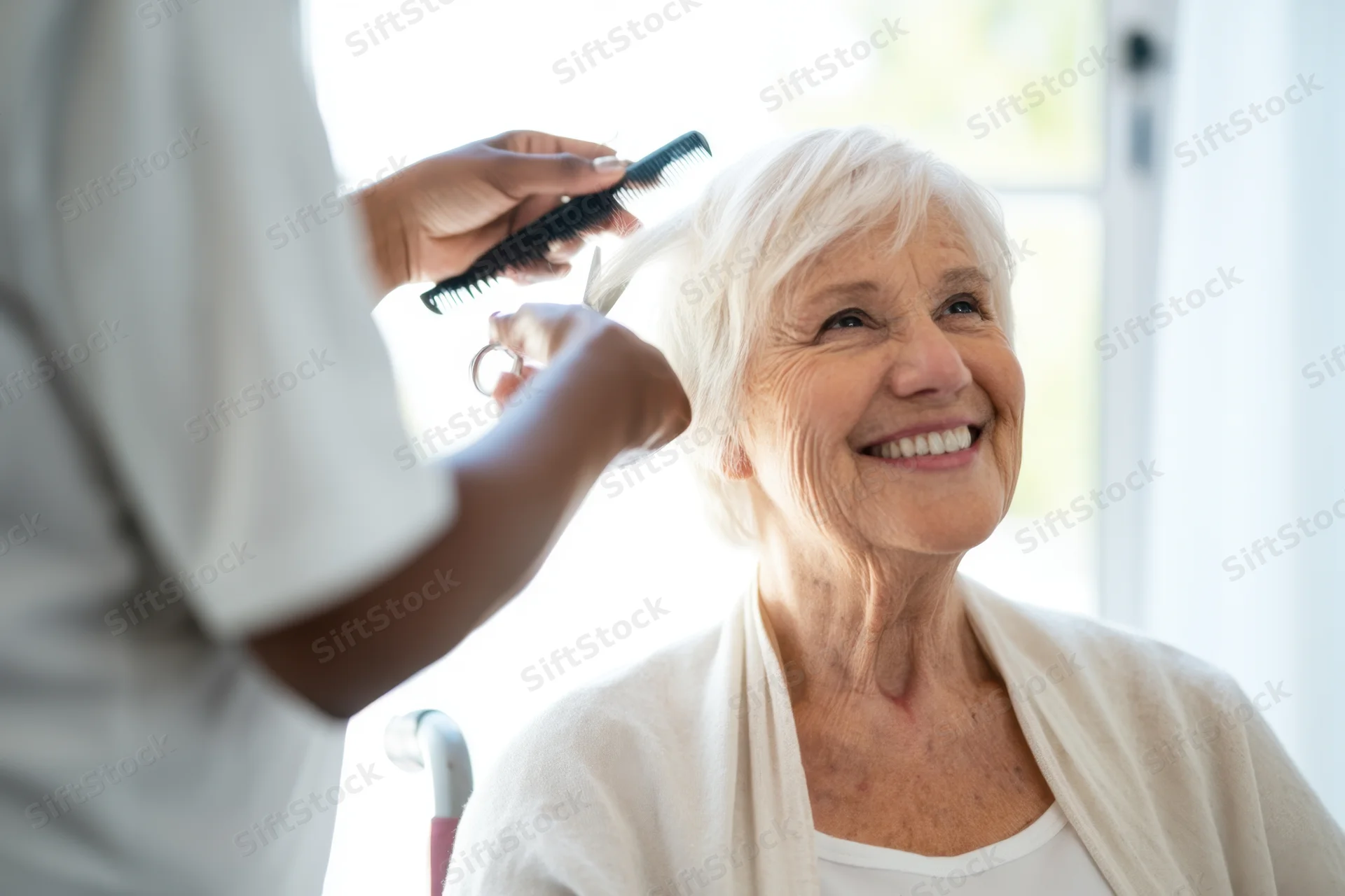Elderly woman smiling while receiving a haircut from a caregiver indoors by a sunny window