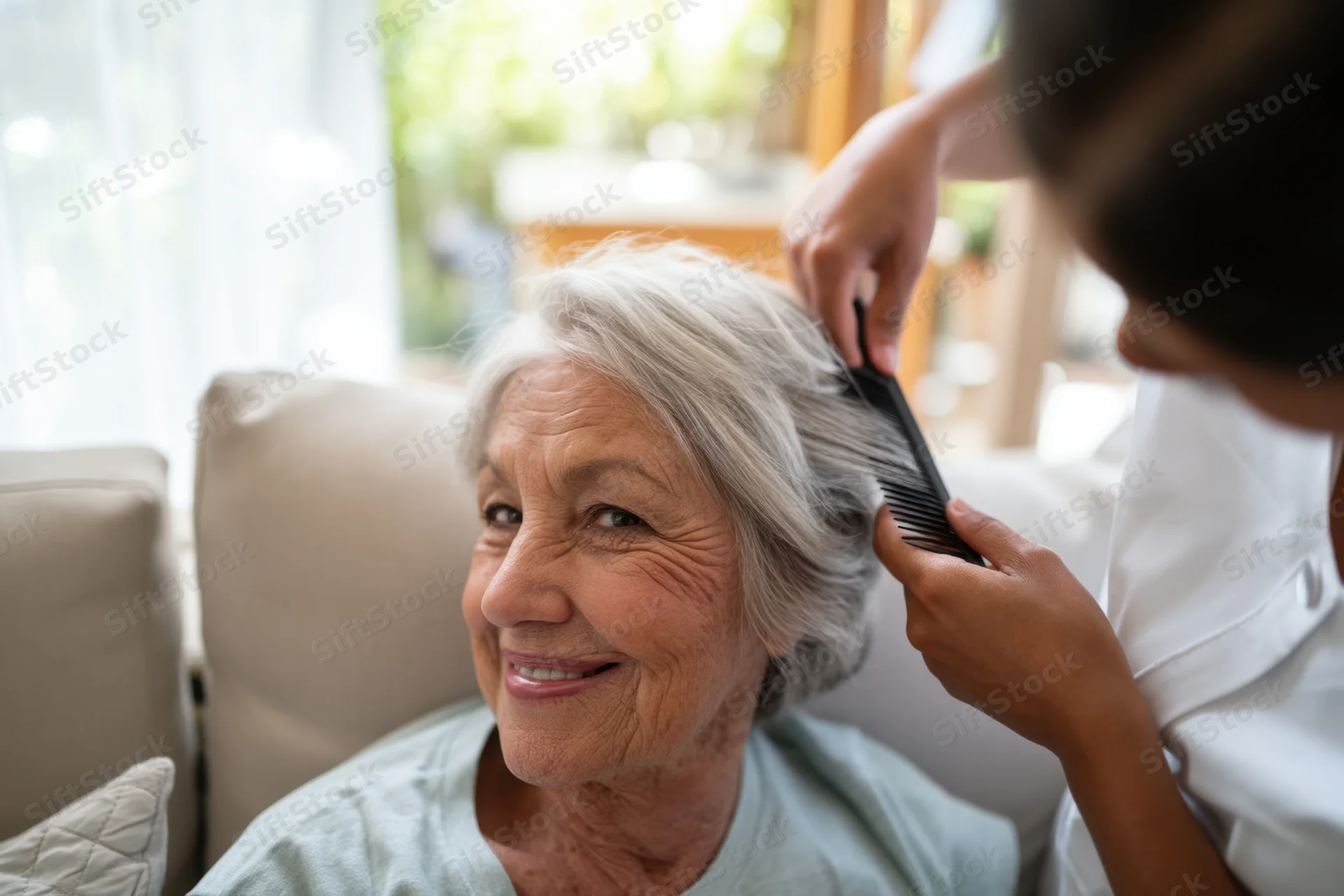 Elderly woman smiling while caregiver combs her silver hair on a sofa in a bright living room