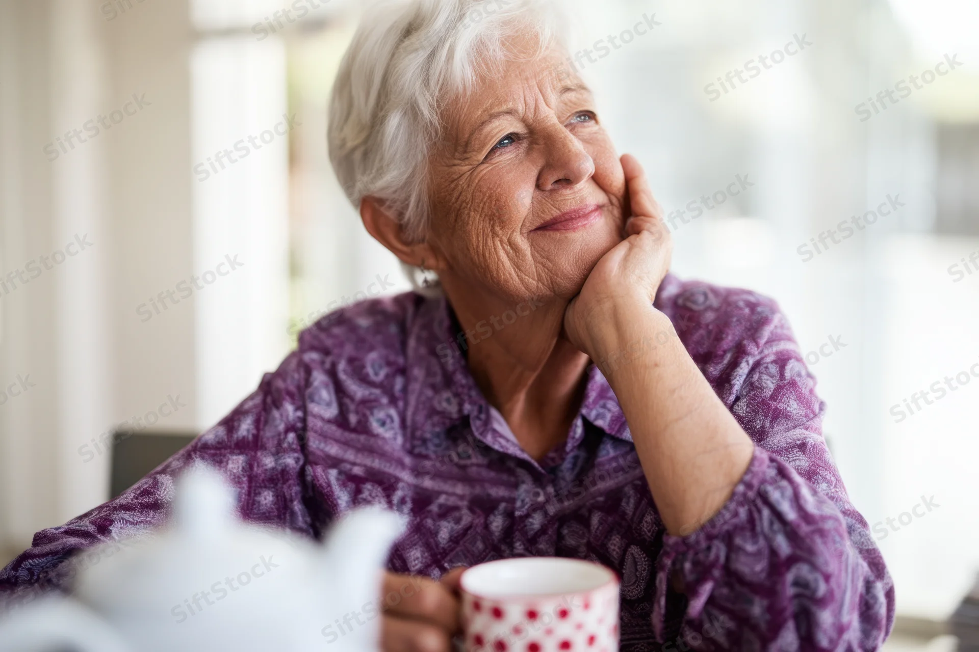 Elderly woman smiling with hand on chin while holding a tea cup at kitchen table