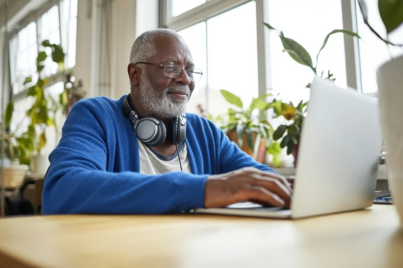 Senior man typing on laptop at wooden desk by window with plants
