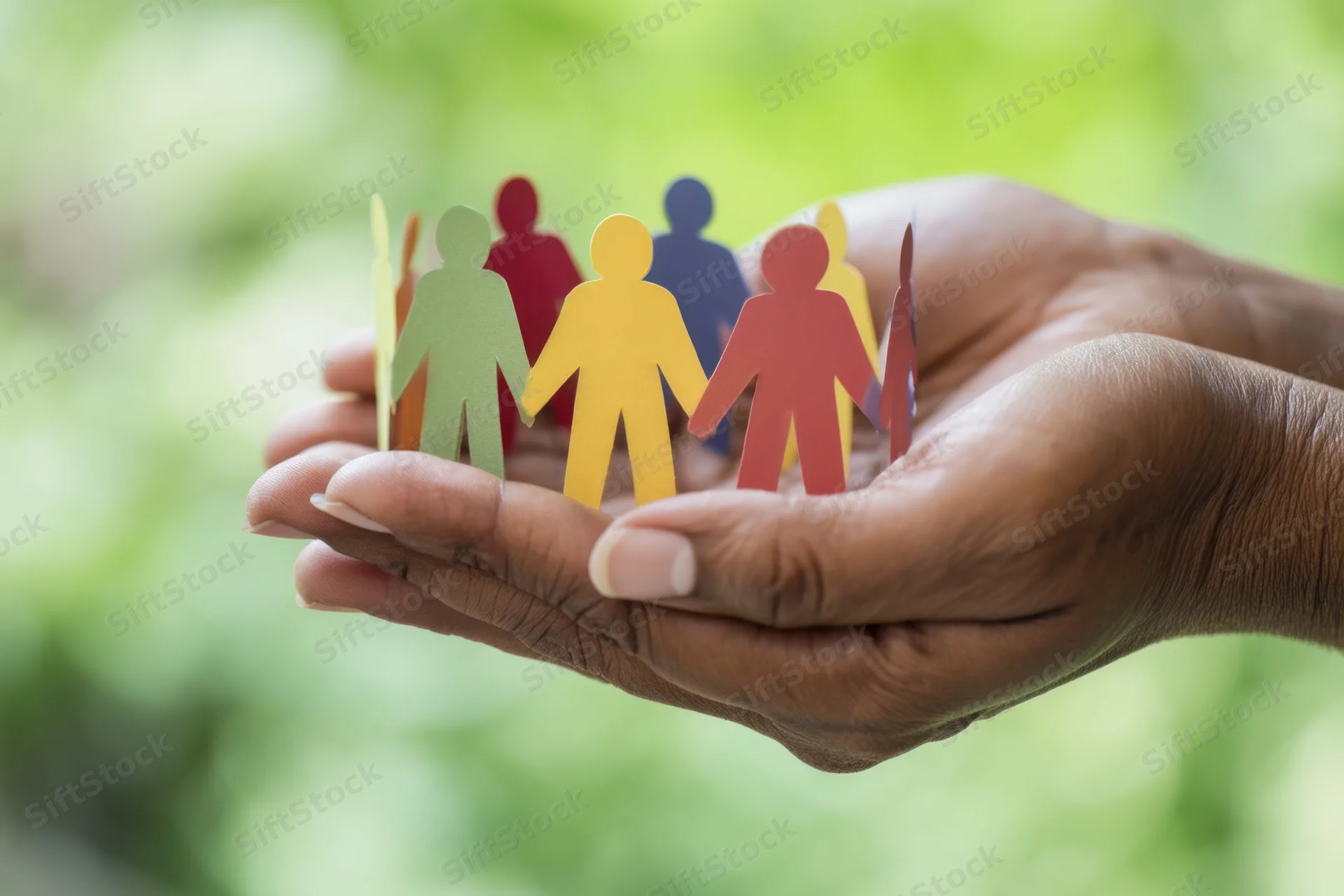 Hands cupping colorful paper cutout people against a green natural background