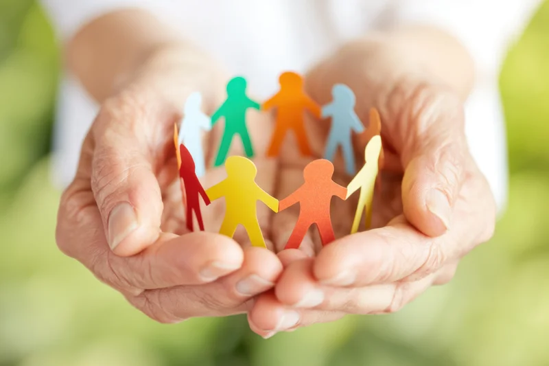 Elderly woman's hands cupping colorful paper cutout people in a circle, outdoors with green background