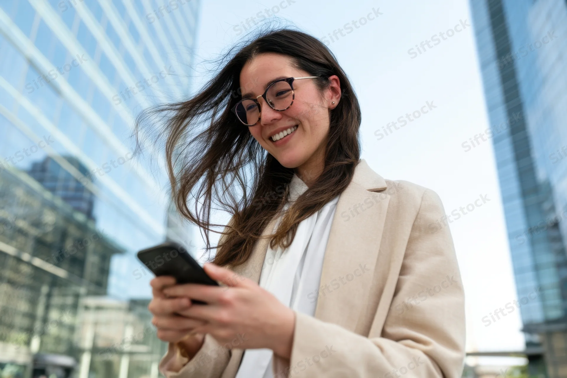 Woman smiling while using smartphone outdoors in city with skyscrapers behind her