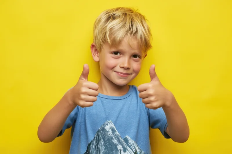 Young blond boy giving two thumbs up against yellow background wearing blue tshirt with mountain print
