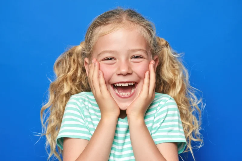 Young girl with blonde pigtails laughing with hands on cheeks against blue background
