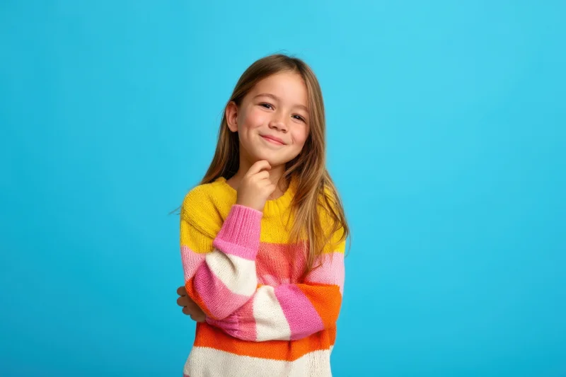 Young girl smiling with hand on chin against blue background wearing colorful striped sweater