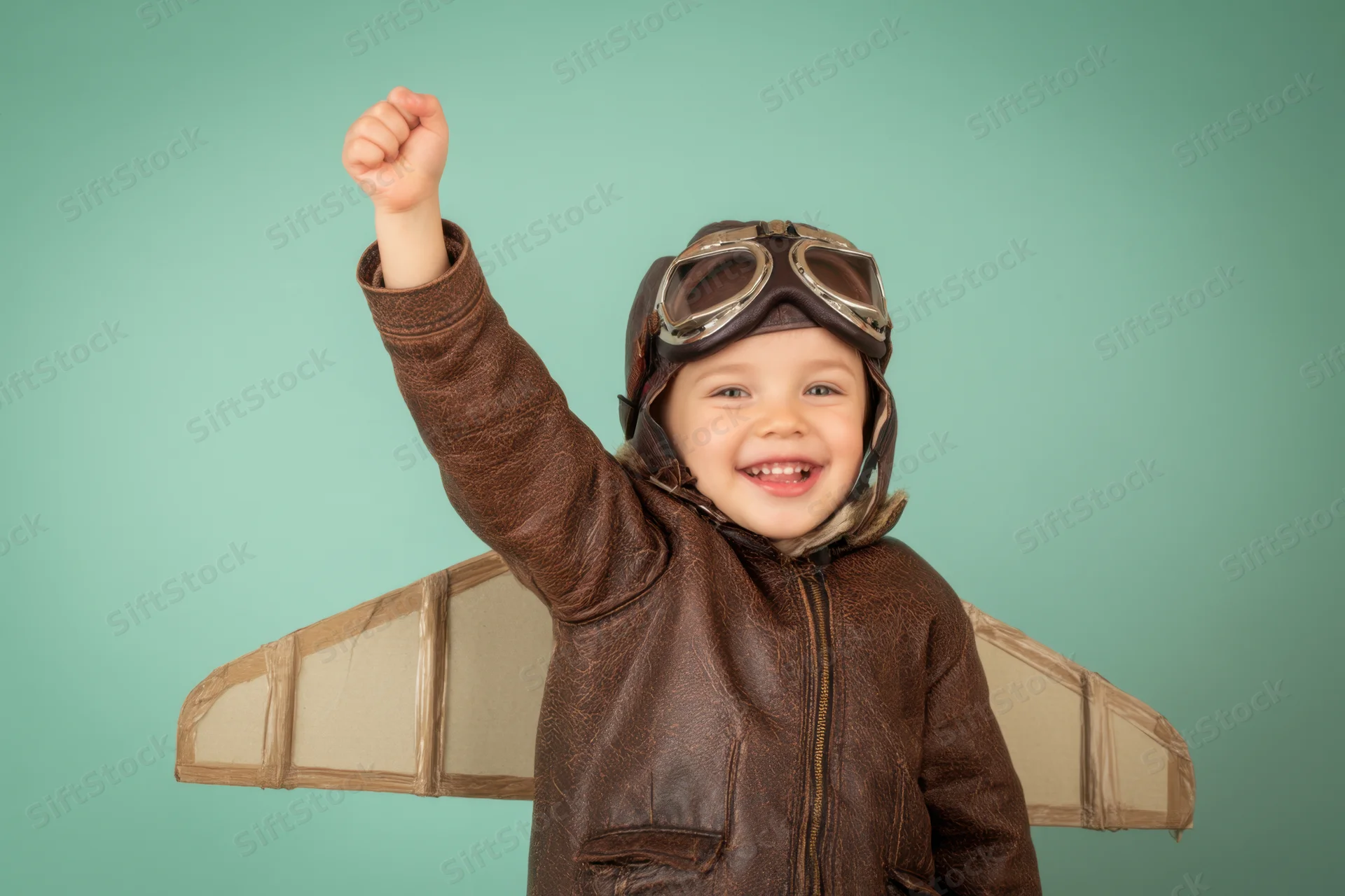 Child in aviator costume with cardboard wings raising fist against teal background