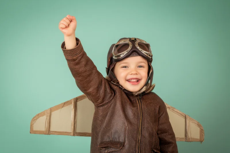 Child in aviator costume with cardboard wings raising fist against teal background