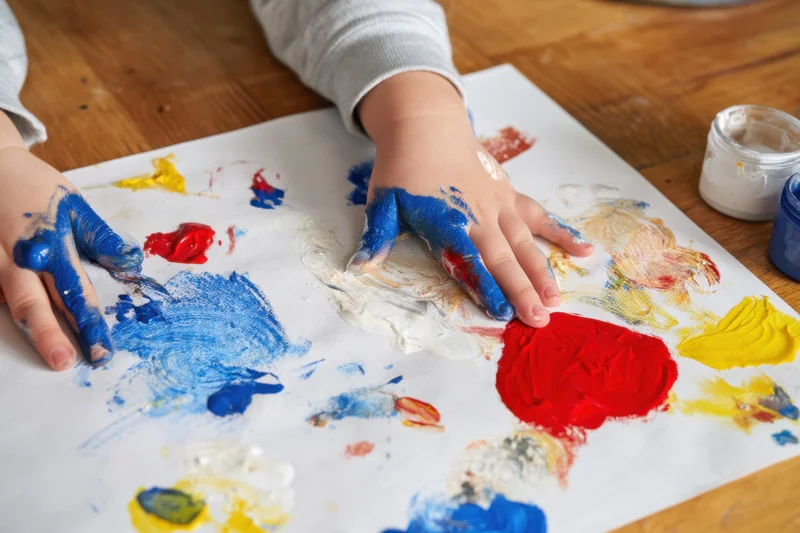 Child's hands covered in blue red yellow and white paint making marks on paper on wooden table