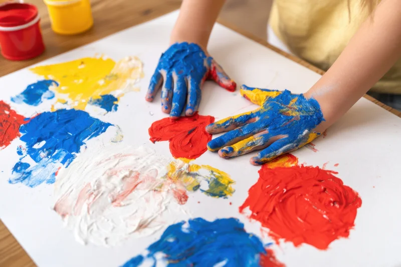 Child’s hands covered in blue red yellow and white paint pressing onto paper on a wooden table