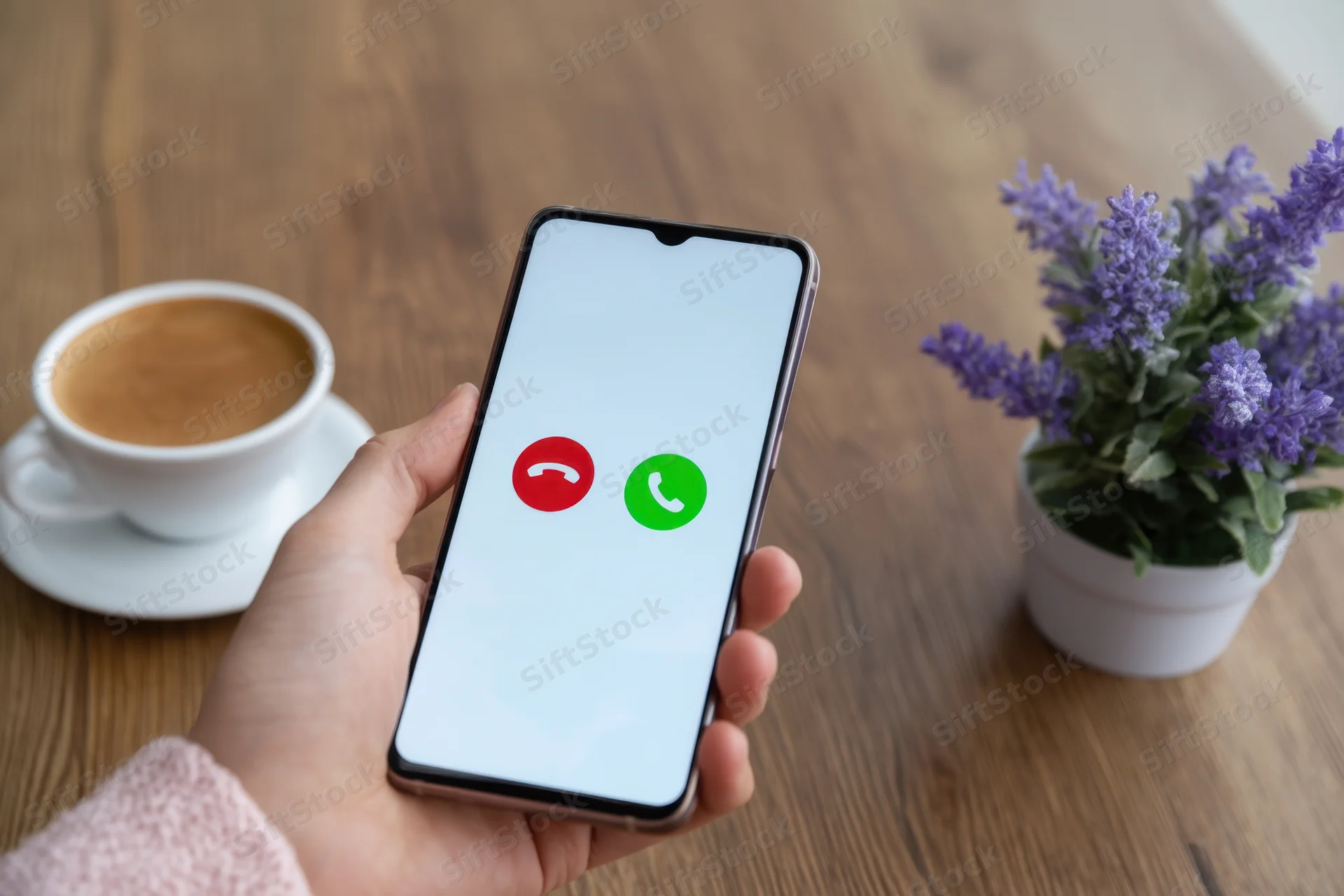 Hand holding smartphone with incoming call screen on wooden table near coffee cup and lavender plant