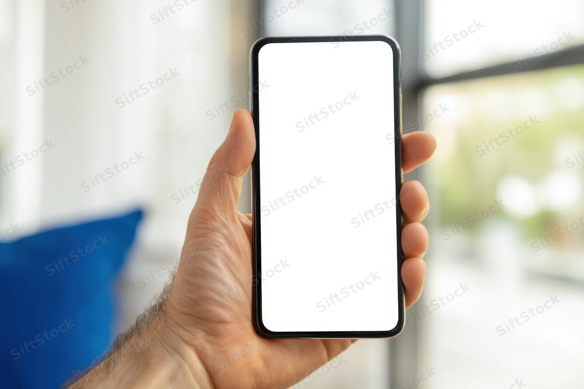 Hand holding smartphone with blank white screen in front of window and blue cushion